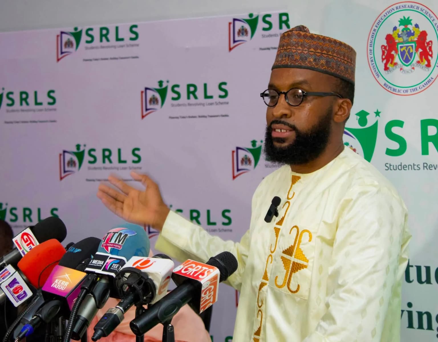 A speaker at an SRLS press briefing, in front of a backdrop carrying the SRLS and Ministry of Higher Education logos, facing a bank of press microphones.