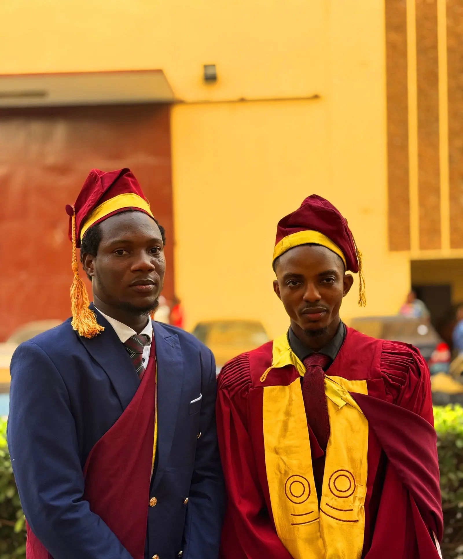 Two graduates in academic gowns and caps standing together outside a building.